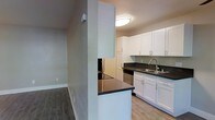 This single-floor space offers a clean, functional layout with modern finishes throughout. The kitchen is well-appointed with crisp white shaker-style cabinetry, dark countertops, and stainless steel appliances, including a dishwasher and an electric cooktop set into a peninsula that separates the kitchen from the adjacent living area. A double-basin sink with a modern faucet sits beneath flush-mount ceiling lighting, lending the kitchen a bright and practical atmosphere. Light-toned flooring runs through the kitchen, contrasting with the darker hardwood-style flooring found in the rest of the unit. The living area is open and airy, featuring neutral-toned walls and large sliding glass doors that flood the space with natural light and provide direct access to an outdoor balcony. Through the glass, mature trees are visible, offering a pleasant, leafy outlook. A wall-mounted HVAC unit provides climate control, and a secondary window further enhances ventilation and natural illumination. The overall design aesthetic is contemporary and understated, with clean lines, neutral tones, and a cohesive material palette that creates a welcoming and versatile living environment.