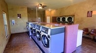 This single-story laundry facility is designed to accommodate high-volume use, featuring a well-organized layout with a row of commercial-grade front-load washing machines positioned along a central island. The machines are mounted on a tiled base and topped with a stainless steel surface, providing a functional workspace. A pink speckled folding counter runs alongside the machines, offering ample space for sorting and folding laundry. Along the back wall, a set of stacked dryers provides additional drying capacity, efficiently maximizing the available floor space. The room is finished with warm, earth-toned textured walls that lend a casual, welcoming ambiance. Terracotta-style tile flooring adds durability and easy maintenance underfoot. A ceiling fan with an integrated light fixture is centrally mounted, ensuring consistent air circulation throughout the space. An electrical panel is also visible, indicating dedicated utility infrastructure for the facility. The overall layout is practical and straightforward, prioritizing functionality while maintaining a clean and organized environment suitable for regular, everyday use.