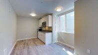 This contemporary single-floor unit offers a clean, efficient layout designed to maximize functionality and comfort. The open-concept living area is finished with light-toned laminate wood flooring throughout, contributing to a bright and cohesive aesthetic. Neutral gray walls and flush-mounted ceiling lighting create a modern, well-lit environment.

The kitchenette is a standout feature, outfitted with sleek white cabinetry — both upper and lower — paired with a dark countertop for contrast. Stainless steel appliances include an over-the-range microwave and a refrigerator, complemented by a built-in cooktop and a single-basin sink. A large window adjacent to the kitchen allows natural light to filter into the space, enhancing its open feel.

The main living area is generously proportioned, with a solid entry door providing privacy and security. A wall-mounted heating unit ensures climate control year-round. The space flows naturally toward the bathroom, which is finished in crisp white tones and features a full shower-tub combination with chrome fixtures. The bathroom also includes a toilet and sink, with accessible grab bar support visible along the walls.

Overall, the unit presents a practical and modern living environment, ideal for those seeking a well-appointed, low-maintenance space with clean finishes and thoughtful design.