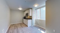 This contemporary single-floor unit offers a clean, efficient layout designed to maximize functionality and comfort. The open-concept living area is finished with light-toned laminate wood flooring throughout, contributing to a bright and cohesive aesthetic. Neutral gray walls and flush-mounted ceiling lighting create a modern, well-lit environment.

The kitchenette is a standout feature, outfitted with sleek white cabinetry — both upper and lower — paired with a dark countertop for contrast. Stainless steel appliances include an over-the-range microwave and a refrigerator, complemented by a built-in cooktop and a single-basin sink. A large window adjacent to the kitchen allows natural light to filter into the space, enhancing its open feel.

The main living area is generously proportioned, with a solid entry door providing privacy and security. A wall-mounted heating unit ensures climate control year-round. The space flows naturally toward the bathroom, which is finished in crisp white tones and features a full shower-tub combination with chrome fixtures. The bathroom also includes a toilet and sink, with accessible grab bar support visible along the walls.

Overall, the unit presents a practical and modern living environment, ideal for those seeking a well-appointed, low-maintenance space with clean finishes and thoughtful design.