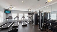This fitness center is a thoughtfully designed, single-floor facility built to support a well-rounded workout routine. The space is finished with durable dark rubber flooring throughout, providing a safe and practical surface for exercise. Light blue-gray walls and white trim contribute to a clean, energizing atmosphere, while multiple large windows fitted with white blinds allow generous natural light to fill the room.

The cardio area is a standout feature, housing a row of treadmills positioned along the windowed wall, accompanied by elliptical trainers and a stationary bike. Wall-mounted flat-screen televisions are positioned for easy viewing during workouts. Ceiling fans with integrated lighting are distributed across the ceiling, ensuring consistent air circulation throughout the space.

A dedicated strength training zone includes a multi-function cable machine and a well-stocked dumbbell rack offering a broad range of free weights, paired with a flat workout bench. Floor-to-ceiling mirrors line one wall, visually expanding the space and allowing users to monitor their form during exercise.

Additional amenities include an informational bulletin board, a paper towel and sanitizing station for equipment upkeep, and convenient access doors. The overall layout is organized and functional, making this fitness center a comprehensive and accessible resource for daily exercise.