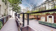 Located at 4601 East Flat Rock Road in Philadelphia's scenic Manayunk neighborhood, this outdoor space is a beautifully landscaped courtyard and deck environment that seamlessly blends urban design with natural elements. The space features multi-level composite decking finished in warm wood tones, connected by a gently sloping accessible walkway with sleek cable and glass railings framed by dark metal posts.

At the heart of the space, a dedicated lounge area offers built-in wood bench seating accented by tiled green bases, accompanied by bistro tables and decorative lanterns. Lush raised planters overflow with trailing greenery and ornamental shrubs, creating a verdant boundary that softens the urban surroundings. Mature trees are integrated throughout the deck, providing natural canopy and dappled shade.

A pergola structure with tall steel columns anchors one end of the space, adding architectural definition and framing an open-air area suitable for gatherings or outdoor dining. String lights are strung overhead, lending an inviting ambiance for evening use. The space is bordered by a classic whitewashed multi-story building alongside a modern gray structure, reflecting the area's blend of historic and contemporary architecture. Together, these elements create a versatile, park-like outdoor retreat in the heart of the city.
