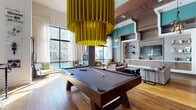 This two-story space makes an immediate impression through its bold, eclectic design language and generous proportions. The entry level welcomes visitors with large-format tile flooring, soaring double-height ceilings, and a striking feature wall housing circular barrel-style booth seating—an inventive architectural focal point that anchors the communal dining area. A long, dark wood communal table surrounded by industrial metal chairs occupies the central gathering zone, illuminated by oversized pendant fixtures. Vibrant teal accent walls and large-scale murals contribute energy and visual depth throughout.

The upper level, accessible via an open staircase with sleek cable railings, transitions to warm wide-plank hardwood flooring. Here, floor-to-ceiling windows frame urban courtyard views, flooding the space with natural light. A full-size pool table commands the game lounge beneath a dramatic oversized chartreuse pendant light. An adjacent lounge area features built-in shelving, a flat-screen TV, and comfortable seating arrangements. Exposed wood beam ceilings define a well-appointed kitchen with a lengthy quartz-topped island, warm wood cabinetry, a mosaic tile backsplash, and pendant lighting. Separate lounge nooks with curated furniture and artwork create intimate retreats within the larger open-plan layout.