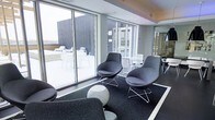 This contemporary single-floor space is designed with a clean, modern aesthetic that emphasizes openness and connectivity between its indoor and outdoor areas. The interior lounge features sleek, rounded bucket-style chairs in charcoal grey with polished chrome wire bases, arranged thoughtfully atop a dark area rug over light flooring — creating a defined and inviting seating zone. Soft stone-toned ottoman accents complement the neutral, sophisticated color palette of grey and white throughout.

Large floor-to-ceiling sliding glass doors form an entire wall of the space, flooding the interior with abundant natural light and offering a seamless visual and physical transition to an outdoor terrace. The terrace is outfitted with white tables and chairs, providing additional seating suited for gatherings or quiet relaxation. The exterior facade visible through the windows features a striking dark vertical-panel cladding, adding architectural contrast to the bright interior.

The ceiling design incorporates subtle recessed lighting, enhancing the polished, professional atmosphere. A mirrored element on one wall extends the sense of depth within the space. Overall, the design strikes a balance between functional comfort and refined, contemporary style, making it well-suited as a communal lounge or hospitality amenity space.