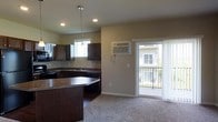 This single-floor space offers a thoughtfully designed open-concept layout that seamlessly connects the kitchen and living area. The kitchen is outfitted with rich, dark wood cabinetry that extends to the upper walls, providing ample storage alongside a deep, functional countertop. A central kitchen island with a breakfast bar adds both workspace and a casual dining option. Recessed lighting and a sleek pendant light above the island keep the kitchen well-illuminated, while a window above the sink brings in natural light. Appliances are integrated neatly into the cabinetry, contributing to a clean, cohesive look. A wall-mounted air conditioning unit ensures year-round comfort. The adjacent living area features neutral carpet flooring, creating a warm and inviting atmosphere that contrasts nicely with the hard flooring of the kitchen. A sliding glass door dressed with sheer white curtains opens to a private balcony, offering views of the surrounding outdoor area and filling the space with natural light. The overall ambiance is modern and practical, with neutral tones throughout that lend a versatile, move-in-ready feel to the space.