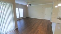 This single-floor residence offers a thoughtfully designed open layout that feels both spacious and welcoming. The main living area is anchored by rich, dark hardwood flooring that runs throughout, providing a warm and polished aesthetic that pairs well with the neutral-toned walls. Crown molding adds a refined architectural detail along the ceiling line, lending the space a classic, elevated character.

A ceiling fan with a light fixture is centrally positioned in the living area, ensuring year-round comfort and functional illumination. Natural light enters through windows fitted with horizontal blinds, brightening the interior and creating an airy atmosphere. The clean white trim along the baseboards and door frames offers a crisp contrast to the warm flooring and soft wall tones.

The space includes a white-paneled interior door leading to an adjacent room, suggesting a well-connected floor plan that flows naturally from one area to the next. The walls are neutral in color, providing a versatile backdrop well-suited for a variety of interior styles and furnishings.

Overall, the space presents a clean, move-in-ready environment with a balance of classic detailing and practical design — ideal for comfortable everyday living.