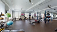 This single-floor fitness center is designed to accommodate a comprehensive range of workout styles within a polished, welcoming environment. The main workout area is generously proportioned, featuring durable rubber flooring throughout and tray ceilings with crown molding that lend an upscale aesthetic uncommon in typical gym settings. Ceiling fans are positioned throughout to ensure comfortable airflow during workouts.

Natural light fills the space through an abundance of white-trimmed windows fitted with horizontal blinds, offering outdoor views while maintaining a bright and energizing atmosphere. Multiple wall-mounted flat-screen televisions are strategically placed for viewing during cardio sessions.

The cardio zone is lined with a row of treadmills and elliptical machines from the Promaxima brand, positioned along the windowed walls. A central functional training area features a blue multi-station power rack system equipped for suspension training, boxing, and barbell work, complemented by plyo boxes and benches. A dedicated free weights section houses a full rack of dumbbells in graduated sizes, while a separate strength zone includes cable machines and resistance equipment.

A connected corridor with a warm wood-panel ceiling leads to a hydration station and storage area, adding convenience. A bright corner room with panoramic windows provides a quieter zone for stretching, stability ball training, and targeted machine work, completing this well-rounded fitness facility.