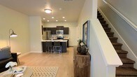 This two-story residence offers a thoughtfully designed layout that balances modern aesthetics with everyday functionality. The main level features an open-concept living and kitchen area, creating a seamless flow ideal for both entertaining and daily living. The kitchen is a standout focal point, outfitted with dark cabinetry that contrasts elegantly against light countertops and a full suite of stainless steel appliances, including a refrigerator, range, and microwave. A central island with bar-height seating provides additional workspace and a casual dining option. Recessed lighting throughout the main level ensures the space is well-lit, complementing the warm, light-toned hardwood floors that run continuously across the open floor plan. Neutral wall tones enhance the bright and airy ambiance of the interior. A carpeted staircase visible from the main living area leads to the second floor, suggesting additional private living quarters above. Near the entry, a rustic console table adds a charming, organic touch that balances the otherwise contemporary design. The overall aesthetic combines clean lines, practical finishes, and warm accents to create a welcoming and functional living environment across both levels.