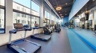Located at 700 Semmes Ave in Richmond, Virginia, this well-appointed fitness center offers a spacious, single-floor layout designed to support a full range of workout routines. The facility is characterized by soaring ceilings with an exposed industrial aesthetic, featuring visible ductwork and bold pendant ring lighting that adds a contemporary flair to the space.

Floor-to-ceiling windows line the perimeter, flooding the interior with abundant natural light and providing open views of the surrounding Richmond streetscape. The flooring throughout is durable rubber gym matting in a speckled charcoal and blue finish, suitable for high-traffic athletic use. Vivid blue accent walls complement the modern design palette and add energy to the environment.

The cardio zone is well-equipped with treadmills, elliptical trainers, stationary bikes, and rowing machines positioned along the windowed wall to maximize the connection to the outdoors. The strength training area features a comprehensive selection of free weights including a full dumbbell rack, weight benches, cable machines, and Life Fitness strength equipment. A dedicated functional training zone includes medicine balls and additional benches, all reflected in large wall-mounted mirrors fitted with mounted flat-screen TVs. The space also benefits from direct exterior access through glass-panel doors, enhancing both ventilation and accessibility.