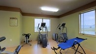 This single-floor fitness room offers a practical and functional space designed for exercise and physical wellness. The room features light-toned walls accented with a warm brown border near the ceiling, contributing to a clean and energizing atmosphere. Durable light wood-grain flooring lines the entire space, providing both a polished look and a sturdy surface suitable for workout activities.

The room is equipped with a selection of cardio and exercise machines, including two elliptical trainers, a stair climber, a stationary cycle, and an adjustable exercise bench, offering users a well-rounded workout experience. A centered window along one wall allows natural light to filter into the space and provides an outdoor view, enhancing the overall environment during workouts.

Overhead fluorescent lighting ensures the room remains well-lit at all times, supporting safe and effective use of the equipment. The layout is thoughtfully arranged to provide ample space between machines, allowing for comfortable movement throughout the room. Overall, this fitness space is a well-appointed and inviting facility for those looking to maintain an active lifestyle.