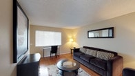 This single-floor space offers a thoughtfully arranged living area that balances comfort and functionality. The living room is anchored by warm hardwood flooring that runs throughout, adding a sense of warmth and continuity to the space. A large window fitted with horizontal blinds allows generous natural light to fill the room, creating a bright and welcoming atmosphere. The living area features a plush dark sofa accented with patterned throw pillows, paired with a sleek round glass-top coffee table resting on a decorative area rug. A wall-mounted mirror adds depth and dimension to the room. A dedicated workspace is neatly integrated into the living area, complete with a desk and chair positioned near the window — ideal for productivity in a well-lit setting. Ambient lighting is provided by a classic floor lamp, contributing to a warm and inviting ambiance during evening hours. The textured ceiling adds a subtle architectural detail throughout the space. Overall, this residence offers a practical and comfortable layout that makes efficient use of its single-floor footprint, with classic finishes and tasteful furnishings that create a cohesive and livable environment.