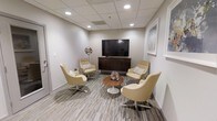This lounge is a thoughtfully designed single-floor space that combines comfort and functionality in a clean, contemporary setting. The room features neutral-toned walls paired with a drop ceiling outfitted with recessed lighting, which creates an even, welcoming illumination throughout the space. The flooring consists of a textured, multi-toned carpet that adds visual interest while contributing to the room's comfortable atmosphere.

At the center of the seating arrangement, four cream-colored swivel chairs with polished chrome bases are positioned around a round pedestal side table, accented with a decorative starburst centerpiece. The chairs are styled with patterned throw pillows, enhancing the inviting nature of the space. A large flat-screen television is mounted above a dark espresso wood media credenza along one wall, serving as a focal point for entertainment or presentations. Decorative accents, including a sculptural sphere and a framed wall print, add personality without overwhelming the space. Overall, the lounge offers a polished, relaxed environment suitable for casual conversation, informal meetings, or quiet downtime.