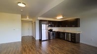This single-floor space showcases a contemporary open-concept layout that seamlessly integrates the kitchen and living areas. The kitchen is a standout feature, equipped with a full suite of stainless steel appliances, including a refrigerator, electric range, microwave, and dishwasher. Rich, dark espresso-toned cabinetry lines the walls, providing generous storage space, while light-colored countertops create a striking contrast against the bold cabinetry. A single-basin sink is positioned along the counter, complemented by under-cabinet task lighting that enhances functionality.

The flooring throughout the space consists of warm, dark hardwood planks that add depth and a sense of continuity across the open layout. Flush-mount ceiling light fixtures provide consistent, even illumination throughout. A hallway visible in the background leads toward the entry door, suggesting a well-organized floor plan. The smooth, neutral-toned walls and clean architectural lines contribute to a minimalist, modern aesthetic. The open sightlines between the kitchen and surrounding areas make the space feel expansive and welcoming, ideal for everyday living and entertaining.