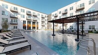 Located at 4600 Ross Ave in Dallas, Texas, this thoughtfully designed outdoor amenity space serves as a vibrant communal hub within a modern multi-story residential community. The centerpiece is a generously sized resort-style swimming pool with crystal-clear water, surrounded by a wide pool deck lined with sleek chaise lounges — ideal for sunbathing beneath the Texas sky. Twin steel pergolas provide shaded gathering areas, furnished with contemporary lounge seating and side tables, creating comfortable spots for relaxation and socializing. Adjacent to the pool, a well-maintained turf lawn offers flexible open space, anchored by a full-size outdoor ping pong table and accented with large decorative planters overflowing with seasonal flowers and shrubs. A quieter seating nook tucks neatly against the building's facade, furnished with plush white lounge chairs and a small bistro table — perfect for a more intimate outdoor retreat. The surrounding landscaping features neatly arranged garden beds with low-maintenance plantings, ornamental trees, and manicured greenery that soften the clean lines of the building's contemporary white and charcoal exterior. Iron railings and balconies from the floors above overlook the courtyard, creating a cohesive, enclosed feel that balances openness with a sense of community.