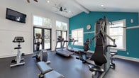 This single-story fitness center is designed to offer a comprehensive workout experience in a bright, welcoming environment. The main exercise area is characterized by vaulted ceilings with dual ceiling fans that promote airflow throughout the space. A bold teal accent wall adds a dynamic visual contrast to the otherwise crisp white walls and trim, creating an energizing atmosphere conducive to exercise.

Large glass-paned doors and generous windows line one side of the room, flooding the interior with natural light and providing views of the surrounding outdoor area, which appears to include a pool. A wall of mirrors on one end visually expands the space and assists with form during workouts.

The facility is well-appointed with a variety of cardio equipment, including treadmills, an elliptical, a stationary bike, and a rowing machine. Strength training is supported by a multi-function cable machine, adjustable benches, a dumbbell rack with a full range of free weights, and kettlebells. A wall-mounted television, water fountain, and water cooler add convenience for users.

A separate corridor provides access to an ADA-accessible restroom. The dark rubber flooring throughout the exercise area offers both durability and safety, completing this fully functional fitness facility.
