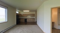 This single-floor space features an open-concept layout that seamlessly connects the kitchen and living area, creating a functional and cohesive environment. The kitchen is outfitted with light wood cabinetry spanning both upper and lower sections, providing generous storage throughout. A continuous countertop with a granite-style laminate finish runs the length of the kitchen wall, incorporating a stainless steel double-basin sink. Black appliances, including a dishwasher and refrigerator, offer a contrast to the warm tones of the cabinetry. The kitchen flooring is a dark hardwood-style surface, adding warmth and durability underfoot. The adjoining living area transitions to wall-to-wall carpeting in a neutral tone, lending a comfortable feel to the space. Flush-mounted fluorescent ceiling fixtures provide bright, even overhead lighting throughout the kitchen and living area. The textured ceiling and neutral-toned walls create a clean, understated backdrop throughout. A baseboard heating unit is visible along the living area wall, and a window allows natural light to enter the space, contributing to an open and airy atmosphere overall.