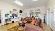 This single-story community clubhouse is thoughtfully designed to serve as a central hub for residents, offering a comprehensive range of amenities across several well-appointed spaces. The welcoming reception and lounge area sets a warm, inviting tone with plush, ornate sofas accented by colorful throw pillows, a decorative area rug over tile flooring, and a wall-mounted television. Large windows allow natural light to fill the space, while a staffed front desk is conveniently positioned nearby. An adjacent seating nook with high vaulted ceilings and large windows overlooking the community provides a quiet retreat. The business center is equipped with desktop computers, a printer, and ample counter space for literature and resources. A casual dining area features multiple table-and-chair sets beneath bright overhead lighting, with windows offering views of the outdoor pool area. The fitness room is outfitted with a multi-station cable machine, exercise bench, stability balls, and a treadmill, set against a warm accent wall with carpeted flooring. A spacious laundry facility houses rows of commercial washers and dryers alongside a utility sink. A compact kitchenette with a full-size refrigerator and countertop rounds out the amenities available to residents.