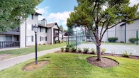 This residential community presents a well-organized, attractively landscaped campus spread across thoughtfully maintained grounds. The multi-story buildings feature classic exterior design elements, including light-colored lap siding, gabled rooflines, brick foundation accents, and dark shutters flanking grid-pane windows that lend a traditional aesthetic throughout the property. Residents benefit from private balconies and patios, offering personal outdoor spaces with views of the surrounding greenery.

The grounds are a standout feature of this community, with lush, manicured lawns, mature deciduous and evergreen trees, and carefully tended garden beds filled with shrubs and seasonal flowering plants in decorative planters. Wide concrete walkways and driveways wind through the property, providing easy pedestrian access between buildings and amenities. Classic lamp-post lighting lines the paths, adding both charm and practicality.

A fenced tennis court is prominently situated within the community, offering recreational opportunities for residents. The overall ambiance is that of a peaceful, park-like setting that balances community living with access to open outdoor spaces. The single-story clubhouse or leasing office building features large grid windows, brick base detailing, and black trim accents, serving as an inviting focal point within the community's layout.