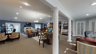 This single-floor facility offers a thoughtfully designed community space that blends classic architectural character with a warm, inviting atmosphere. The centerpiece of the space is a generously sized open lounge area, defined by elegant white decorative columns with ornate capitals that frame the room and add a touch of traditional sophistication. Vaulted ceilings with a chandelier and recessed lighting enhance the sense of openness, while soft blue-gray walls create a calm, welcoming ambiance throughout.

The lounge is furnished with plush brown sofas and accent chairs accented by vibrant teal, mustard, and orange throw pillows, arranged around coffee tables to encourage conversation and community gathering. A fireplace with a white mantel serves as a focal point at one end of the lounge, complemented by a wall-mounted television.

Adjacent to the lounge is a functional kitchenette featuring warm oak cabinetry, dark countertops, a double stainless steel sink, and hardwood flooring — providing a practical space for refreshments and everyday use.

A dedicated office area is also present, equipped with a large desk, visitor seating, and wall-mounted display shelves showcasing framed awards and credentials. Natural light filters in through multiple windows, further brightening the overall space and contributing to its professional yet comfortable character.