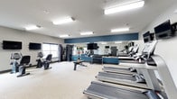 This fitness center offers a comprehensive workout environment designed to accommodate a wide range of exercise routines. The space is finished with light carpet flooring and illuminated by rows of bright fluorescent overhead lighting, creating a clean and energizing atmosphere throughout.

Along one side of the room, a row of treadmills provides ample cardio options, complemented by a stationary recumbent bike and additional cardio equipment nearby. A full-length mirror spans a prominent accent wall painted in a deep navy blue, visually expanding the space and allowing users to monitor their form during exercise.

The free weights area features a well-organized dumbbell rack stocked with a wide selection of weights, alongside colorful medicine balls for functional training. A weight bench is centrally positioned for versatile strength exercises. A multi-function cable and strength training machine is available along the perimeter, offering a variety of resistance training options.

Mounted televisions are positioned throughout the room, ensuring entertainment is accessible from multiple workout stations. Windows along the walls allow natural light to supplement the overhead lighting, contributing to an open and welcoming environment. The overall layout is thoughtfully arranged to maximize usability and flow, making this fitness center a fully functional and well-appointed space for daily exercise.
