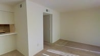 This single-story space features a clean, minimalist interior with neutral cream-colored walls that create a bright and open atmosphere. The room is finished with wall-to-wall carpeting in a light beige tone, contributing to a warm and comfortable feel underfoot. Natural light filters into the space, casting defined streaks across the floor and highlighting the simplicity of the layout. A ceiling-mounted HVAC vent ensures climate control throughout. A doorway leads to an adjacent closet, offering practical built-in storage. The door features simple, functional hardware consistent with the understated design aesthetic of the space. Electrical outlets are positioned along the baseboards, providing convenient access for furnishings and devices. The walls are smooth and uniform, providing a neutral backdrop well-suited for a variety of décor styles. The overall ambiance is calm and uncluttered, making the space highly adaptable. The straightforward single-floor layout promotes ease of movement and accessibility, while the consistent use of neutral tones throughout lends the space a cohesive, move-in-ready character.