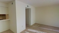 This single-story space features a clean, minimalist interior with neutral cream-colored walls that create a bright and open atmosphere. The room is finished with wall-to-wall carpeting in a light beige tone, contributing to a warm and comfortable feel underfoot. Natural light filters into the space, casting defined streaks across the floor and highlighting the simplicity of the layout. A ceiling-mounted HVAC vent ensures climate control throughout. A doorway leads to an adjacent closet, offering practical built-in storage. The door features simple, functional hardware consistent with the understated design aesthetic of the space. Electrical outlets are positioned along the baseboards, providing convenient access for furnishings and devices. The walls are smooth and uniform, providing a neutral backdrop well-suited for a variety of décor styles. The overall ambiance is calm and uncluttered, making the space highly adaptable. The straightforward single-floor layout promotes ease of movement and accessibility, while the consistent use of neutral tones throughout lends the space a cohesive, move-in-ready character.