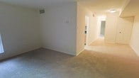 This single-story space is designed with a straightforward, functional layout that connects its main living areas through a central hallway. The living area features wall-to-wall neutral-toned carpet, which extends throughout the space, creating a consistent, cohesive feel underfoot. Smooth, cream-colored walls are kept clean and minimal, providing a blank canvas adaptable to a variety of interior styles.

Overhead HVAC vents are positioned throughout, indicating a central air and heating system for year-round climate control. A partial wall divides the main living area from a corridor that leads deeper into the unit, where a glimpse of warm wood-toned cabinetry suggests a kitchen with practical storage. The hallway continues further, with additional rooms visible in the background, likely including one or more bedrooms and a bathroom, as indicated by the carpeted flooring and door frames visible from the main area.

The space is well-lit with ceiling-mounted lighting visible along the hallway. The overall ambiance is clean, understated, and move-in ready, suited for those looking for a simple, neutral environment.