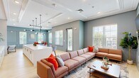 Located at 700 City View Loop in Midlothian, Virginia, this thoughtfully designed single-floor amenity space offers a versatile and welcoming environment for gathering, socializing, and collaboration. The open-concept layout connects multiple functional zones, including a spacious lounge area, a kitchen bar, and dedicated meeting and seating nooks, all unified by a cohesive aesthetic of cool blue-gray walls, crisp white crown molding, and light hardwood flooring.

The lounge area is anchored by a large sectional sofa and complemented by modern swivel chairs arranged beneath a wall-mounted television, flanked by elegant panel molding and soft sconce lighting. Adjacent to the lounge, a central kitchen bar features a sleek white countertop, terracotta subway tile backsplash, and a contemporary multi-arm pendant light fixture, with bar seating that invites casual conversation.

A dedicated meeting room offers a round dark-wood table surrounded by bold orange upholstered chairs, bathed in natural light from multiple large windows with outdoor views. Additional seating alcoves provide quieter retreat spaces. Throughout, recessed lighting, decorative wall paneling, and tray ceilings with detailed crown molding reinforce the space's polished, modern-classic character.