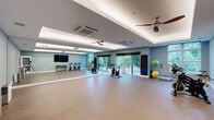 This dedicated fitness studio offers a generously sized, open floor plan designed to accommodate a variety of exercise activities. The space is anchored by a striking tray ceiling with built-in ambient lighting that casts a warm, even glow throughout the room, complemented by two ceiling fans that ensure comfortable airflow during workouts. Durable, cushioned cork-style flooring covers the entire studio, providing both comfort and support underfoot.

A full-wall mirror spans one side of the room, enhancing the sense of openness while allowing users to monitor their form during exercise. The studio is equipped with multiple stationary cycling bikes arranged for group or individual use, along with stability balls, resistance accessories, and a punching bag for a well-rounded fitness experience. A wall-mounted flat-screen television adds a multimedia element to the space.

Soft blue walls lend a calm, focused atmosphere to the room. Large sliding glass doors and windows along one wall flood the interior with natural light and provide views of a lush outdoor balcony area, creating an energizing connection to the outdoors. Ceiling-mounted HVAC units ensure year-round climate control. A secondary entrance with glass-paneled doors offers convenient access, rounding out this thoughtfully designed, fully functional fitness environment.