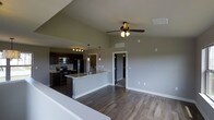 This two-floor space showcases a thoughtfully designed open-concept layout, seamlessly connecting the kitchen and living areas into a cohesive, functional environment. The kitchen is outfitted with dark espresso cabinetry that creates a striking contrast against light granite countertops. Stainless steel appliances, including a refrigerator, range, and microwave, add a sleek, modern touch. A peninsula with an integrated sink extends into the main living space, offering additional counter space and casual seating potential. Pendant lighting above the peninsula complements a dedicated track lighting fixture over the main kitchen area, providing layered illumination. The living area features dark hardwood-style flooring that flows continuously throughout the open space, grounding the neutral gray wall palette. A ceiling fan with a built-in light kit is centrally positioned, enhancing airflow and ambient lighting. HVAC vents are discreetly integrated into the ceiling, maintaining the clean aesthetic. A doorway visible from the main space provides access to additional rooms, and the two-floor layout offers further versatility and living space beyond the main level.