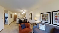 This single-floor residence offers an open-concept layout that seamlessly connects the living, dining, and kitchen areas into one cohesive and functional space. The living area is comfortably appointed, featuring a plush seating arrangement with a warm rust-toned armchair and a blue sofa, complemented by decorative wall art and a stylish floor lamp that adds a welcoming ambiance. A ceiling fan overhead provides both light and air circulation throughout the space. Adjacent to the living area, the dining space accommodates a round table with seating for multiple guests, making it well-suited for everyday meals and casual entertaining. The kitchen is positioned just beyond, featuring a peninsula-style breakfast bar, white cabinetry, and standard appliances, offering a practical and efficient cooking environment. Neutral-toned carpeting flows throughout the living and dining areas, contributing to the overall understated and clean aesthetic. The open sightlines between zones make the space feel airy and connected, while the straightforward, uncluttered design ensures ease of movement and livability throughout.
