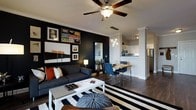 This single-floor space showcases a confident modern aesthetic with an open-concept layout that seamlessly connects the living, dining, and kitchen areas. The living room makes a strong visual statement with a dramatic black accent wall adorned with a curated gallery of framed artwork, providing a bold backdrop to the charcoal sofa accented with rust-colored throw pillows. A bold black-and-white striped area rug anchors the seating area beneath a classic ceiling fan, while a clean white coffee table adds contrast and balance. A pendant cluster chandelier elegantly defines the dining zone, where a sleek wooden dining table is paired with modern navy chairs. The open kitchen features white cabinetry, stainless steel appliances, and a breakfast bar pass-through, all illuminated by recessed track lighting. Decorative column details add an architectural touch at the transition between spaces. Dark hardwood flooring flows continuously throughout, tying the entire layout together with warmth and sophistication. The overall ambiance is urban and polished, blending contemporary design elements with curated décor for a cohesive and inviting atmosphere.