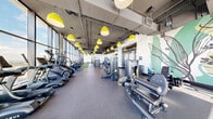 This expansive fitness center offers a generously sized, single-floor layout designed to accommodate a full range of workout disciplines. The space is defined by dramatic floor-to-ceiling glass windows that wrap along the perimeter, flooding the interior with abundant natural light and providing sweeping panoramic views of the surrounding cityscape and waterways — an inspiring backdrop for any workout.

The industrial-modern aesthetic is carried throughout, with exposed concrete ceilings, visible ductwork, and bold yellow pendant lights suspended in a row overhead, adding a striking pop of color against the neutral palette. Durable rubber flooring covers the main exercise areas, while a turf lane inset into the floor provides a dedicated space for functional training drills and agility work.

Equipment is thoughtfully organized across the floor, with a full lineup of premium cardio machines — including treadmills, StairMasters, ellipticals, and stationary bikes — positioned directly facing the expansive windows. The strength training zone features a comprehensive selection of Hoist and Precor cable machines, plate-loaded equipment, and a free weights area stocked with dumbbells and medicine balls. A punching bag station adds further versatility. A large-scale botanical mural accent wall adds an artistic, energizing touch to the overall ambiance.