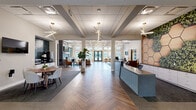 Located at 1340 Wilburn Blvd. in Orbetz, Ohio, this single-story community clubhouse presents a thoughtfully designed interior that balances contemporary aesthetics with comfortable, functional spaces. The entry lobby immediately establishes the building's refined character, featuring a herringbone-patterned hardwood floor, a sleek reception desk, and a striking accent wall composed of hexagonal wood panels set against a lush vertical green installation. Geometric sputnik-style light fixtures illuminate the space with warm, directional light.

Moving deeper into the clubhouse, the main lounge opens into a generously proportioned gathering area with coffered ceilings accented by wood inlays and bold linear pendant lighting arranged in angular configurations. This space includes multiple seating arrangements, a centrally positioned billiards table set atop a patterned blue area rug, and a wall-mounted television above a sleek linear gas fireplace framed in dark tile. Expansive windows and French doors along the perimeter flood the interior with natural light while offering views of the outdoor amenity areas.

A dedicated café nook features a round dining table, a dark tiled accent wall, and a built-in beverage and snack station with a mini-refrigerator and microwave. Throughout, warm hardwood flooring, exposed brick accents, and lush indoor plants contribute to an inviting, upscale ambiance.