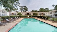 Knollwood is a residential community that offers a welcoming outdoor environment designed for relaxation and recreation. The centerpiece of the community's common area is a generously sized swimming pool with clear turquoise water, framed by a brick-edged pool deck. A collection of lounge chairs lines the perimeter, providing ample space for residents to unwind outdoors.

The surrounding landscape is lush, with tall, mature pine and deciduous trees creating a natural canopy that adds privacy and a sense of seclusion. Well-maintained hedgerows and greenery further enhance the grounds, giving the outdoor space a tidy, park-like feel.

The residential buildings visible from the pool area are multi-story structures clad in neutral tan siding with brown trim and pitched roofs. Each unit appears to feature a private balcony or patio, offering personal outdoor space with views of the landscaped grounds. The overall aesthetic of the community is understated and functional, combining practical amenities with natural surroundings to create a comfortable living environment at Knollwood.