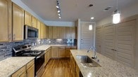 This single-floor space showcases a thoughtfully designed kitchen that balances functionality with modern aesthetics. The kitchen is anchored by warm honey-toned wood cabinetry that extends from floor to ceiling, providing generous storage throughout. A striking mosaic tile backsplash in neutral tones of gray and beige adds texture and visual interest along the walls, complementing the rich hardwood flooring beneath.

The centerpiece of the kitchen is a spacious island topped with light-speckled granite countertops, featuring a deep stainless steel undermount sink paired with a tall gooseneck faucet — ideal for both food preparation and cleanup. Matching granite countertops continue along the perimeter, offering ample workspace.

Stainless steel appliances, including a range and an over-the-range microwave, lend a contemporary edge to the space. Overhead, a multi-head track lighting system provides targeted illumination across the work areas, while a sleek pendant light hangs above the island, adding a warm, focused glow. The kitchen opens toward a well-lit entryway, featuring a solid paneled door and additional closet access, contributing to an open and welcoming flow throughout the space.