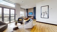 The Sky Lounge in Charlotte, NC (28206) is a sophisticated 1,030-square-foot communal amenity space designed with modern elegance. The single-floor layout seamlessly blends a relaxed lounge area with a fully equipped bar and kitchen zone. The lounge features soaring ceilings, expansive floor-to-ceiling windows and glass doors that flood the space with natural light while offering panoramic views of the surrounding Charlotte skyline and cityscape. A wall-mounted fireplace set against a textured, dark accent wall serves as a focal point, flanked by a mix of neutral and bold-toned seating arrangements on wood-look flooring. The kitchen and bar area is a standout feature, anchored by a long, dramatic marble waterfall island with bar seating. Dark cabinetry, a white subway tile backsplash, open display shelving, a wall-mounted television, and a dedicated wine refrigerator complete the space. Overhead, a striking cluster of mixed-metal pendant lights suspended from an orange-accented ceiling adds a bold design statement. Throughout the space, curated artwork, layered textiles, and carefully chosen furnishings contribute to an upscale yet inviting ambiance, ideal for social gatherings and events.