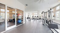 This spacious fitness center is designed to support a full range of workout activities within a clean, modern environment. The interior is finished with neutral white walls and smooth gray flooring throughout, creating a bright and functional atmosphere. Large windows with horizontal blinds line multiple walls, allowing generous amounts of natural light to fill the space and offering views of palm trees and a pool area beyond. Clerestory windows positioned above the primary window line further enhance the sense of openness and contribute to the airy quality of the room. A ceiling fan is centrally mounted, providing additional air circulation across the main workout floor.

The main area is generously proportioned and houses a well-rounded selection of equipment, including treadmills, a StairMaster, an elliptical, a rowing machine, and multiple cable and strength training machines. A separate enclosed room, accessible via glass sliding doors, is dedicated to flexibility and functional training, equipped with stability balls, resistance tools, and TRX suspension straps. Together, these spaces offer a comprehensive and well-organized fitness environment suited to a variety of training styles and fitness levels.