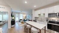 Located in Mauldin, South Carolina, this single-floor unit offers a well-designed layout that maximizes functionality and style. The open-concept living and kitchen area serves as the heart of the space, where light wood-tone flooring, recessed lighting, and a ceiling fan create a bright, welcoming atmosphere. Natural light flows in through large windows and a glass door that opens to the outdoors.

The kitchen is outfitted with crisp white cabinetry, a subtle tile backsplash, granite-style countertops, and a full suite of stainless steel appliances, including a refrigerator, range, microwave, and dishwasher. A central kitchen island with a white countertop and bar-height seating provides additional prep space and a casual dining option.

The living area offers a comfortable, open setting with room for seating and entertainment. Two bedrooms feature wood-look flooring, ceiling fans, and ample natural light from well-placed windows, providing restful retreats with a clean, modern aesthetic.

The full bathroom includes white cabinetry, a granite-top vanity, a large mirror, and a shower-tub combination. A dedicated laundry room houses a full-size washer and dryer with overhead wire shelving for added storage. The overall design balances practicality with contemporary style throughout.
