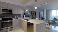 This well-appointed single-floor space showcases a seamless open-concept layout that connects the kitchen and dining areas, creating a cohesive and inviting living environment. The modern kitchen is a standout feature, boasting light wood-toned cabinetry that lends warmth against cool grey walls. A clean white countertop extends across the kitchen island, which houses an undermount sink and integrated dishwasher, making it both functional and visually refined. A textured mosaic tile backsplash adds depth and character to the cooking area. Overhead, two industrial-style pendant lights hang above the island, complementing the recessed track lighting that illuminates the space with a bright, ambient glow. The open layout flows naturally into the dining area, where a curated display of artwork and decorative accessories—including a striking blue canvas painting and an elegant candelabra arrangement—adds a touch of sophistication. A dark wood bookshelf serves as both a storage solution and a design accent, contributing to the layered, gallery-like aesthetic of the space. The overall ambiance is contemporary and curated, blending functionality with modern design sensibilities to create a space that feels both livable and visually engaging.