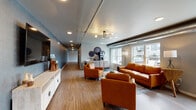 This single-floor space presents a thoughtfully designed communal lounge area that blends contemporary industrial aesthetics with inviting, residential comfort. The main lounge is anchored by a cohesive seating arrangement of rich cognac leather sofas and accent chairs, centered around a round wooden coffee table atop a patterned area rug. Exposed metallic ductwork runs overhead alongside a sputnik-style chandelier, adding an industrial-chic character to the otherwise warm and welcoming atmosphere. Large windows along one wall flood the space with generous natural light, offering views of the surrounding exterior. Warm wood-tone luxury vinyl plank flooring extends throughout, contributing to the cohesive, modern feel. A white credenza along the feature wall provides additional storage and display space. Adjacent to the lounge, a dedicated beverage station with a mini-fridge and counter space offers added convenience for occupants. The space also includes an elevator lobby and secondary access corridor, finished with neutral gray walls, a drop-panel ceiling, and clean, functional flooring. Together, these areas form a well-appointed, multi-functional common space suited for relaxation and community gathering.