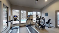 This fitness center is a thoughtfully designed, single-level space offering a comprehensive workout environment. Large windows line the walls, flooding the room with natural light while providing views of the surrounding pool area and landscaped grounds. The neutral-toned walls, carpeted flooring, and ceiling fans contribute to a clean, comfortable atmosphere conducive to exercise.

The cardio zone is well-stocked with multiple True-brand treadmills, elliptical trainers, and a recumbent bike, all positioned to take advantage of the outdoor views. A wall-mounted flat-screen television provides entertainment during workouts. The strength training area features a multi-station cable machine and various weight benches, with floor-to-ceiling mirrors along one wall that enhance the sense of space and allow users to monitor their form.

Access points include a keyed exterior door leading directly to the outdoor grounds and a set of glass double doors that open into an adjacent lounge area, creating a seamless transition between the fitness space and the communal living area. Posted fitness studio rules ensure a respectful environment for all users. The overall design balances functionality with an inviting, light-filled ambiance suitable for a range of fitness routines.