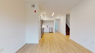 This well-designed two-floor space presents a clean, modern aesthetic defined by light hardwood flooring, crisp white walls, and recessed lighting throughout. The main level features an open layout where the living area flows seamlessly into a contemporary kitchen. The kitchen is equipped with stainless steel appliances—including a range, microwave, refrigerator, and dishwasher—complemented by a two-tone cabinet design pairing white lower cabinetry with dark upper cabinets, and a matte black faucet at the sink. A staircase with dark wood treads connects the two floors, adding a contrasting visual element to the otherwise light interior. The upper level hosts a hallway that leads to additional rooms, including a bright bedroom with large windows fitted with roller shades, allowing ample natural light. The bathroom features large-format travertine-style tile on both the floors and walls, a soaking tub with shower, and matte black fixtures throughout, lending a refined, contemporary character. The overall ambiance of the space is clean, minimal, and move-in ready, with thoughtful finishes and a cohesive modern design language across both floors.