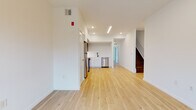 This well-designed two-floor space presents a clean, modern aesthetic defined by light hardwood flooring, crisp white walls, and recessed lighting throughout. The main level features an open layout where the living area flows seamlessly into a contemporary kitchen. The kitchen is equipped with stainless steel appliances—including a range, microwave, refrigerator, and dishwasher—complemented by a two-tone cabinet design pairing white lower cabinetry with dark upper cabinets, and a matte black faucet at the sink. A staircase with dark wood treads connects the two floors, adding a contrasting visual element to the otherwise light interior. The upper level hosts a hallway that leads to additional rooms, including a bright bedroom with large windows fitted with roller shades, allowing ample natural light. The bathroom features large-format travertine-style tile on both the floors and walls, a soaking tub with shower, and matte black fixtures throughout, lending a refined, contemporary character. The overall ambiance of the space is clean, minimal, and move-in ready, with thoughtful finishes and a cohesive modern design language across both floors.