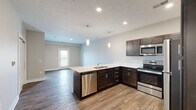 This single-story space is designed with a modern, open-concept layout that seamlessly connects the kitchen and living areas. The kitchen is a standout feature, equipped with dark espresso cabinetry, crisp white countertops, and a full suite of stainless steel appliances, including an electric range, over-the-range microwave, refrigerator, and dishwasher. Pendant lighting hangs above the kitchen peninsula, which doubles as a functional workspace and casual dining area. Recessed lighting further brightens the space.

Wood-style plank flooring runs throughout the main living areas, adding warmth and a cohesive aesthetic. The living room benefits from access to a door leading to a private outdoor area and is bathed in natural light. A bedroom features plush carpeting and a large window offering outdoor views of the surrounding landscape, creating a calm and comfortable retreat.

The bathroom maintains the same design consistency found throughout, with dark cabinetry, a vanity with a white countertop, a mirror, and wood-style flooring. Neutral wall tones and clean white trim are present throughout the entire unit, contributing to a bright, contemporary ambiance that feels both polished and move-in ready.