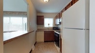 This three-floor home offers a practical and comfortable layout designed for everyday living. The kitchen is a standout functional space, featuring rich dark wood cabinetry that provides ample storage both above and below the countertops. Natural light filters in through a window above the sink, brightening the room and creating a welcoming atmosphere. The kitchen is well-equipped, including a gas range, a dishwasher, a refrigerator, and a microwave, making it fully outfitted for meal preparation. The countertops are neutral in tone, complementing the warm cabinetry, while the light-toned tile flooring adds a clean, classic finish to the space. The layout is efficiently organized in an L-shape, maximizing usability within the available footprint. A secondary window on the adjacent wall further enhances the natural light throughout the kitchen area. With three floors, this home provides distinct zones for living, sleeping, and additional functional use, offering separation and privacy across its levels. The overall ambiance of the kitchen is cozy and utilitarian, with warm wood tones balancing the neutral walls and light fixtures overhead.