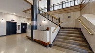 Located in the heart of downtown Saint Paul, this distinguished two-floor commercial building exudes classic architectural grandeur throughout. The expansive lower level serves as an impressive entry point, featuring polished terrazzo floors, bold dark structural columns, and marble-clad surfaces that set a refined tone from the moment of arrival. Elevators framed in rich marble surrounds provide functional connectivity between floors, while a sweeping staircase with ornate wrought-iron railings and brass handrails commands attention as a central architectural feature.

The upper floor is notably generous in scale, offering wide-open corridors adorned with decorative terrazzo tile flooring in geometric patterns and vintage pendant lighting. A striking semi-circular arched window above the main entrance floods the upper landing with natural light, highlighting the intricate coffered ceiling details and elaborate crown moldings found throughout. Warm wood paneling, classic panel doors, and tall ceilings contribute to the building's timeless character.

Accessible via both staircase and glass-enclosed elevator shaft, each level transitions seamlessly, reinforcing the building's blend of historic charm and modern functionality. The overall ambiance reflects a carefully preserved institutional aesthetic, making this a compelling and architecturally significant commercial property in Saint Paul's urban core.