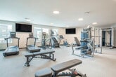 Located at 6604 Odana Rd in Madison, Wisconsin, this well-appointed fitness center offers a generously sized, single-floor layout designed to accommodate a comprehensive range of workout activities. The space is bright and open, with recessed and flush-mount ceiling lighting complemented by natural light streaming through multiple windows that provide views of the surrounding community.

The cardio zone is equipped with a row of Matrix treadmills, stair climbers, and stationary cycling bikes, each paired with individual monitors and wall-mounted flat-screen TVs for entertainment during workouts. A large floor-to-ceiling mirror with a green accent border spans one wall, visually expanding the space and supporting proper form during exercise.

The strength training area includes a multi-station Matrix cable machine, a full dumbbell rack ranging across multiple weight increments, adjustable weight benches, and a rowing machine, offering a thorough free-weight and resistance training experience. The flooring transitions between carpeted and rubberized surfaces suited to different workout zones.

Additional amenities include a dual drinking fountain station, a wall-mounted hand sanitizer dispenser, and a vibrant piece of colorful artwork that adds a touch of energy to the environment. A clearly marked exit door ensures safety and ease of access throughout the facility.