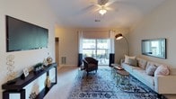 This single-floor residence offers a thoughtfully designed layout with multiple bedrooms, two bathrooms, a functional kitchen, and a comfortable living area. The living room is a welcoming space featuring vaulted ceilings, plush carpeting, and a ceiling fan. A sliding glass door floods the room with natural light and opens to a private balcony, providing outdoor views of surrounding greenery. The room accommodates a tufted sofa, accent chair, and decorative furnishings arranged around a patterned area rug.

The kitchen is efficiently laid out in a galley style, featuring warm wood cabinetry, dark countertops, a double sink, and black appliances including a refrigerator, electric range, and dishwasher. Light tile flooring runs throughout this space.

The bedrooms are carpeted and each includes a ceiling fan, natural light from windows with curtain treatments, and ample room for full-sized furniture. One bedroom features a walk-in closet with built-in shelving. The bathrooms include dark-topped vanities with wood cabinetry and tub-shower combinations. One bathroom features a soaking tub, while the second offers a standard shower enclosure with built-in shelving.