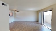 This single-floor space presents a clean, open living area designed for functional simplicity. The room features neutral-toned ceramic tile flooring laid in a grid pattern, providing a durable and easy-to-maintain surface throughout. Smooth, white walls and a plain ceiling create a bright, airy atmosphere, while a ceiling fan with an integrated light fixture offers both ventilation and ambient lighting. A sliding glass door fitted with vertical blinds lines one wall, serving as a key architectural feature that invites natural light deep into the interior while also providing direct access to an outdoor area. The surrounding trim and door frames are finished in white, maintaining a consistent, understated aesthetic. The layout is notably open, offering flexible use of the space with minimal visual obstruction. A window is also visible on an adjacent wall, further enhancing the natural light available. The overall ambiance is neutral and move-in ready, well-suited for a variety of furnishing arrangements and interior styles.